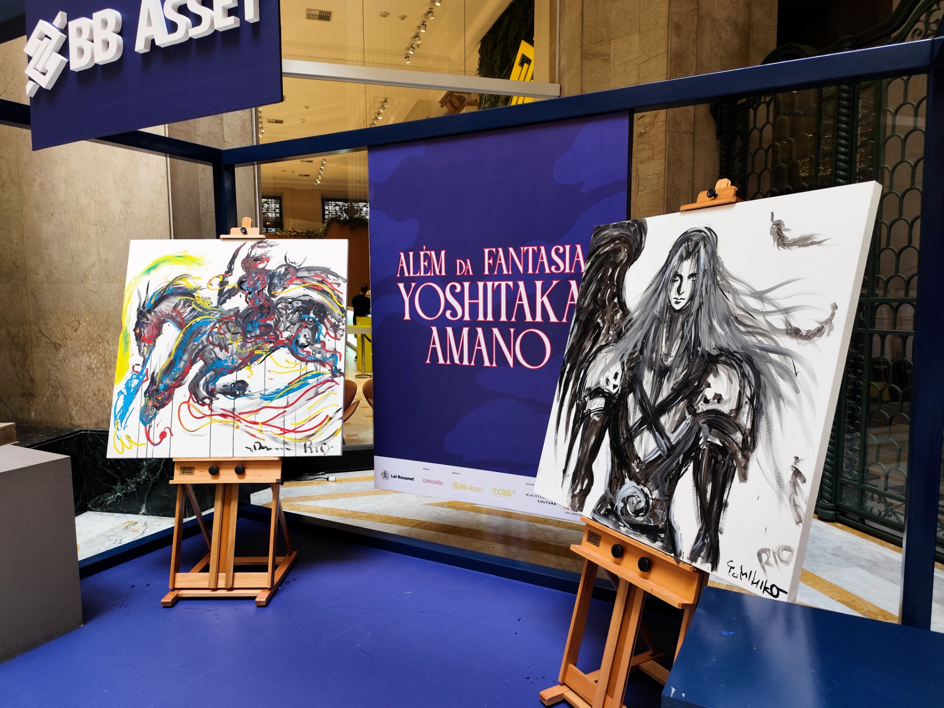 Uma foto de uma exibição de arte no CCBB Rio para a exposição "Além da Fantasia Yoshitaka Amano". Duas pinturas em cavaletes ladeiam um grande banner azul com o título e texto da exposição em português. A pintura da esquerda é colorida, com linhas em azul, amarelo e vermelho. A pintura da direita é monocromática em tons de cinza e preto e apresenta um personagem com longos cabelos brancos e uma asa de anjo preta. O chão é azul brilhante e a estrutura em volta da exibição é preta.