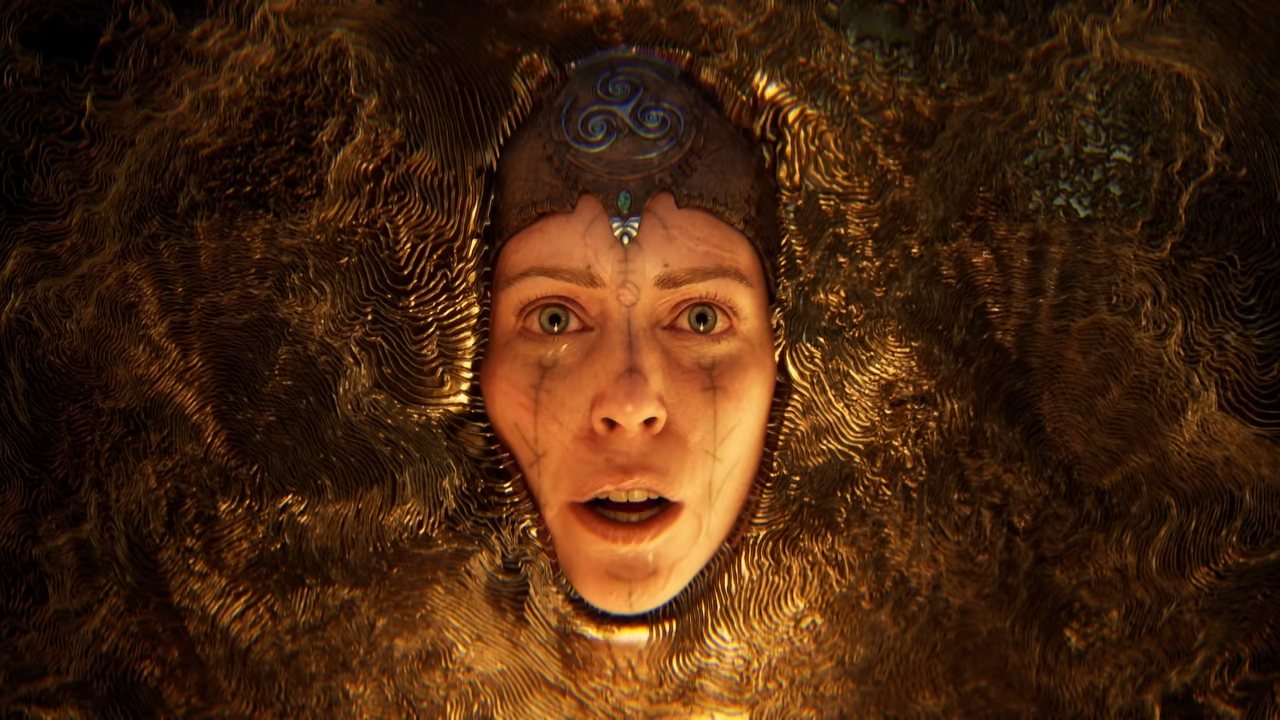 Imagem promocional em grande plano detalhado do rosto da personagem Senua, do jogo "Senua". O rosto expressivo e realista da guerreira celta surge centralizado, emergindo diretamente de uma superfície metálica dourada e texturizada que lembra ouro fundido ou entalhado com finas linhas onduladas. Ela tem olhos claros e arregalados que expressam angústia, choque ou sofrimento, e sua boca está levemente aberta. Linhas de pinturas faciais escuras e desgastadas descem verticalmente por suas bochechas a partir dos olhos, imitando lágrimas. No topo da cabeça, ela usa uma tiara ou adorno de metal escuro. A iluminação é dramática e focada na face, destacando o forte tom dourado e a carga emocional da cena.