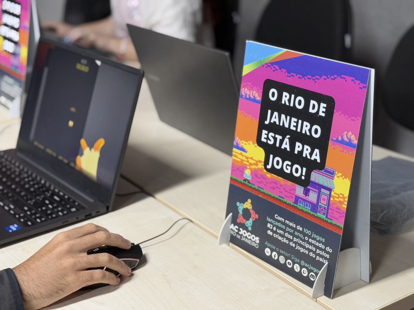 Fotografia em ângulo diagonal de uma bancada de desenvolvimento ou evento de games. No primeiro plano à direita, destaca-se um display de mesa vertical com um cartaz colorido em pixel art nas cores rosa, roxo e laranja. O cartaz exibe o texto em letras maiúsculas: "O RIO DE JANEIRO ESTÁ PRA JOGO!". Abaixo, há um texto menor que diz: "Com mais de 100 jogos lançados por ano, o estado do RJ é um dos principais polos de criação de jogos do país!", acompanhado do logotipo da AC JOGOS (Associação de Desenvolvedores de Jogos Digitais do Rio de Janeiro) e ícones de redes sociais. À esquerda, a mão de uma pessoa segura um mouse gamer preto conectado a um notebook aberto, cujo conteúdo da tela está ligeiramente desfocado. Ao fundo, há outros notebooks e cadeiras de escritório em um ambiente corporativo ou de feira de tecnologia.