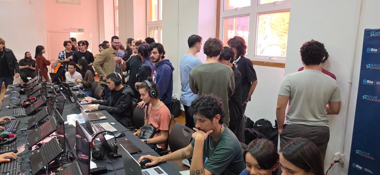 Fotografia em plano aberto mostrando o interior de uma sala movimentada durante um evento de tecnologia ou games na Nave do Conhecimento, no Rio de Janeiro. À esquerda, há uma longa fileira de mesas pretas equipadas com diversos notebooks gamers, fones de ouvido (headsets) com iluminação LED e mouses. Vários jovens estão sentados jogando ou testando softwares; um rapaz de casaco preto usa um headset e mexe no notebook, enquanto ao seu lado outro jovem de camisa salmão também interage com o computador. No centro e ao fundo, dezenas de pessoas, em sua maioria jovens, conversam em pé, criando uma atmosfera de feira de tecnologia ou hackathon. À direita, um banner vertical azul exibe os logotipos da Prefeitura do Rio e do projeto "Nave do Conhecimento - Engenho do Dentro". O ambiente possui janelas grandes ao fundo e iluminação clara.