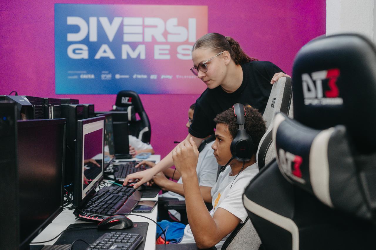 Fotografia em médio plano em uma sala de jogos e tecnologia do projeto "DiversiGames". No centro, um jovem negro de camiseta branca e fones de ouvido (headset) pretos está sentado em uma cadeira gamer cinza e preta da marca DT3, atento à tela de um computador. Ao lado dele, uma jovem de óculos e camiseta preta está inclinada, orientando o aluno e apontando para o teclado e mouse. Ao fundo, a parede exibe um grande painel azul e rosa com o logotipo do projeto "DIVERSI GAMES" e marcas de patrocinadores. Em primeiro plano, no canto direito, destaca-se o encosto de cabeça de outra cadeira gamer preta e branca.