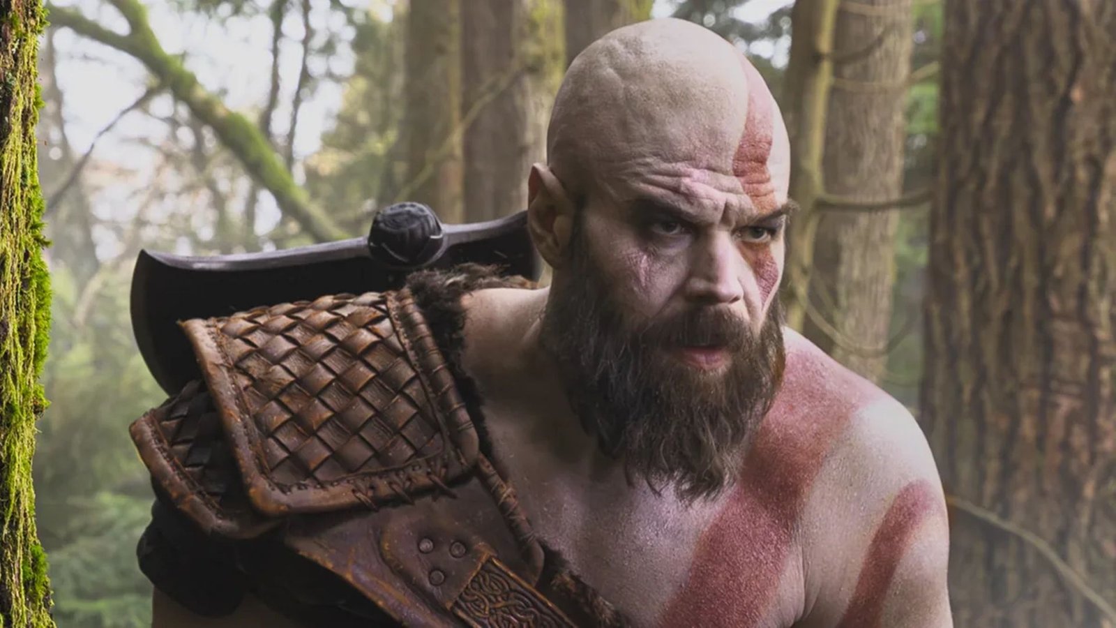 Close de Kratos de God of War, interpretado por Ryan Hurst, em uma floresta nórdica. Imagem mostra a tatuagem vermelha facial de Kratos, barba cheia e armadura de couro com o Machado Leviatã.