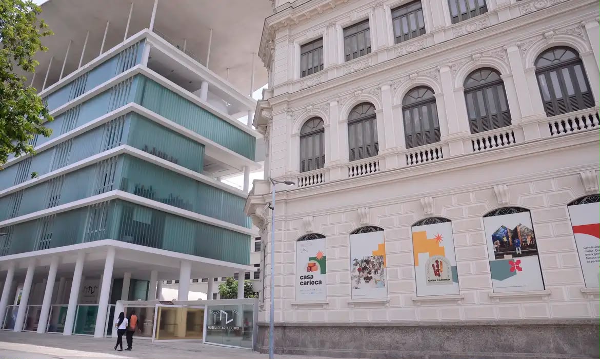 Visão externa do Museu de Arte do Rio (MAR), Rio de Janeiro. A imagem mostra a arquitetura icônica que une o histórico Palácio Dom João VI e o moderno pavilhão de exposições com telhado ondulado. Banners da exposição "Casa Carioca" em destaque. Duas pessoas caminham em frente.