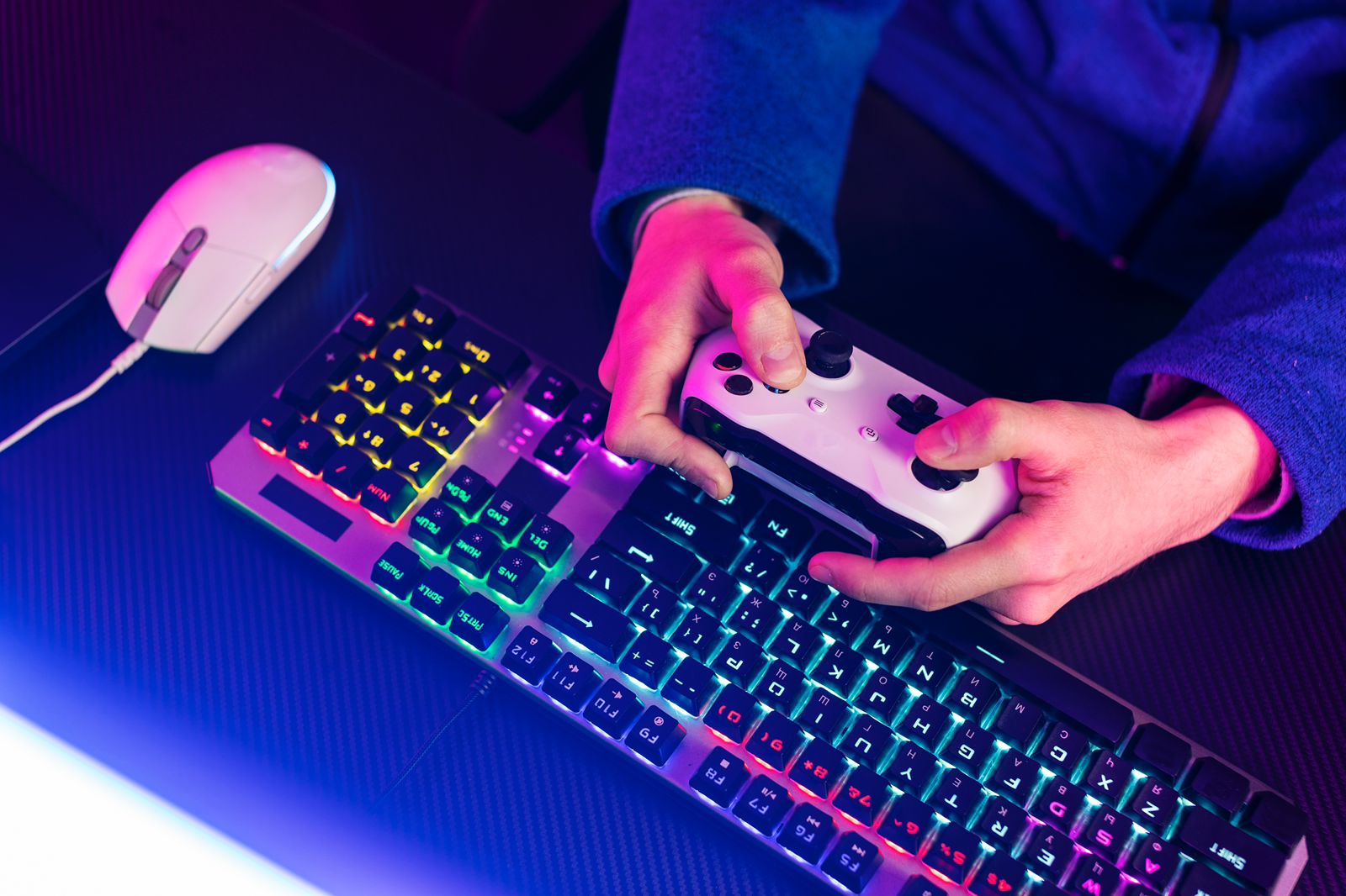 Fotografia em plano picado de um jogador com as mãos em um controle de videogame branco, acima de um teclado mecânico gamer com iluminação RGB azul e rosa em um ambiente escuro.