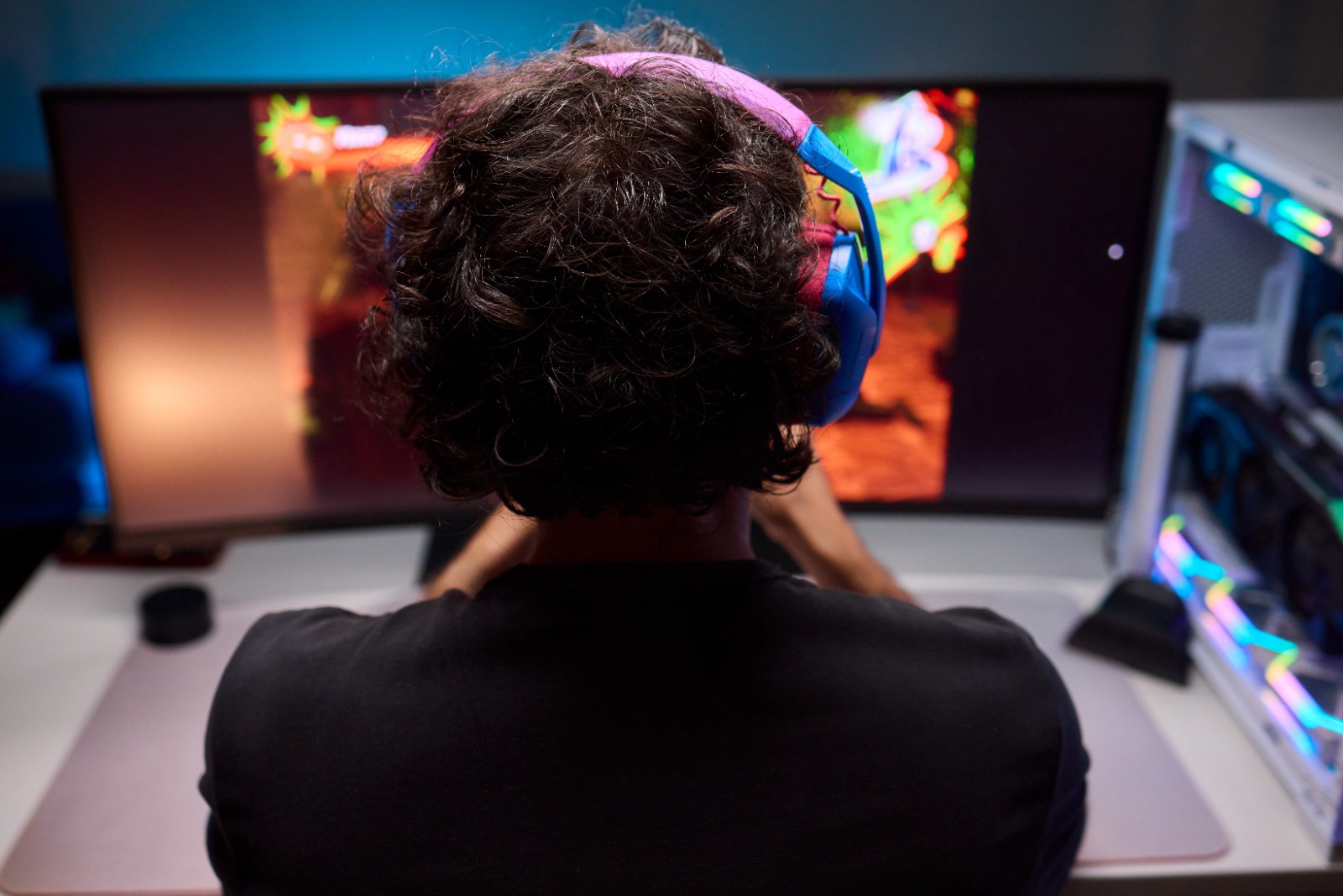 Gamer de costas jogando videogame em um computador de alta performance com monitor ultrawide e fone de ouvido colorido. Foto stock representando setup gamer, streaming e eSports.